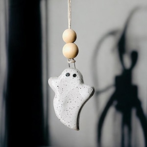 May include: A white ghost-shaped ceramic ornament with black eyes hangs from a string with two wooden beads.