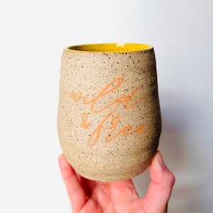 Peut inclure: Une tasse en céramique beige avec un bord jaune et les mots "wild & free" écrits en orange sur le côté.
