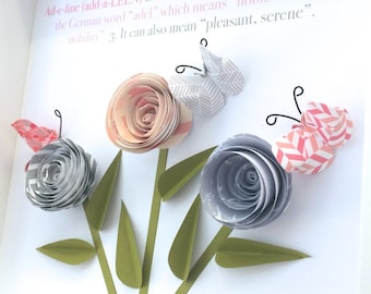 May include: A framed print with the name "Adeline" and a definition of the name's origin and meaning. The print is decorated with three paper flowers and butterflies in shades of pink, gray, and green.