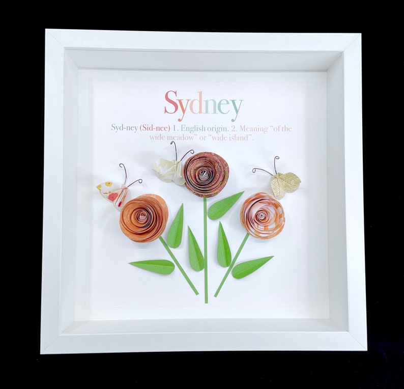 Pode incluir: Uma moldura branca com um passe-partout branco e um fundo branco. A palavra "Sydney" est&aacute; escrita em uma fonte decorativa na parte superior da moldura. Abaixo da palavra "Sydney" h&aacute; uma defini&ccedil;&atilde;o do nome. A defini&ccedil;&atilde;o est&aacute; escrita em texto preto. H&aacute; tr&ecirc;s flores de papel na moldura, cada uma com uma borboleta acima. As flores s&atilde;o laranja e rosa, e as borboletas s&atilde;o brancas, douradas e marrons.