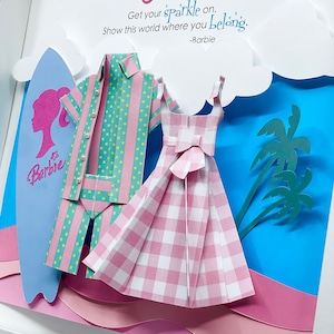 K&ouml;nnte beinhalten: Ein gerahmtes Papierbastelwerk mit einem blauen Surfbrett, einem rosa Kleid, einem blau-rosa gestreiften Hemd und einer Palme. Der Text "Cassie" befindet sich oben im Rahmen. Der Text "Get your sparkle on. Show this world where you belong. -Barbie" befindet sich unter dem Bild.