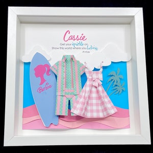K&ouml;nnte beinhalten: Ein wei&szlig;er Rahmen mit einem Papier-Ausschnitt-Design in Blau, Rosa und Gr&uuml;n. Das Design beinhaltet ein Surfbrett, ein Hemd, ein Kleid und Palmen. Der Text "Cassie" befindet sich oben am Rahmen, und der Text "Get your sparkle on. Show this world where you belong. - Rorbie" befindet sich unterhalb des Designs.