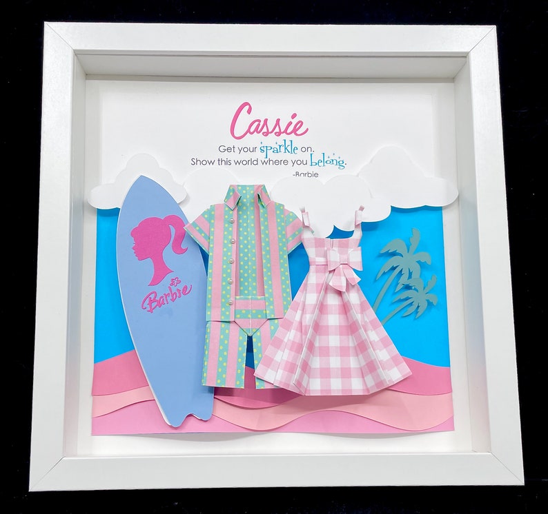 K&ouml;nnte beinhalten: Ein wei&szlig;er Rahmen mit einem Papier-Ausschnitt-Design in Blau, Rosa und Gr&uuml;n. Das Design beinhaltet ein Surfbrett, ein Hemd, ein Kleid und Palmen. Der Text "Cassie" befindet sich oben am Rahmen, und der Text "Get your sparkle on. Show this world where you belong. - Rorbie" befindet sich unterhalb des Designs.