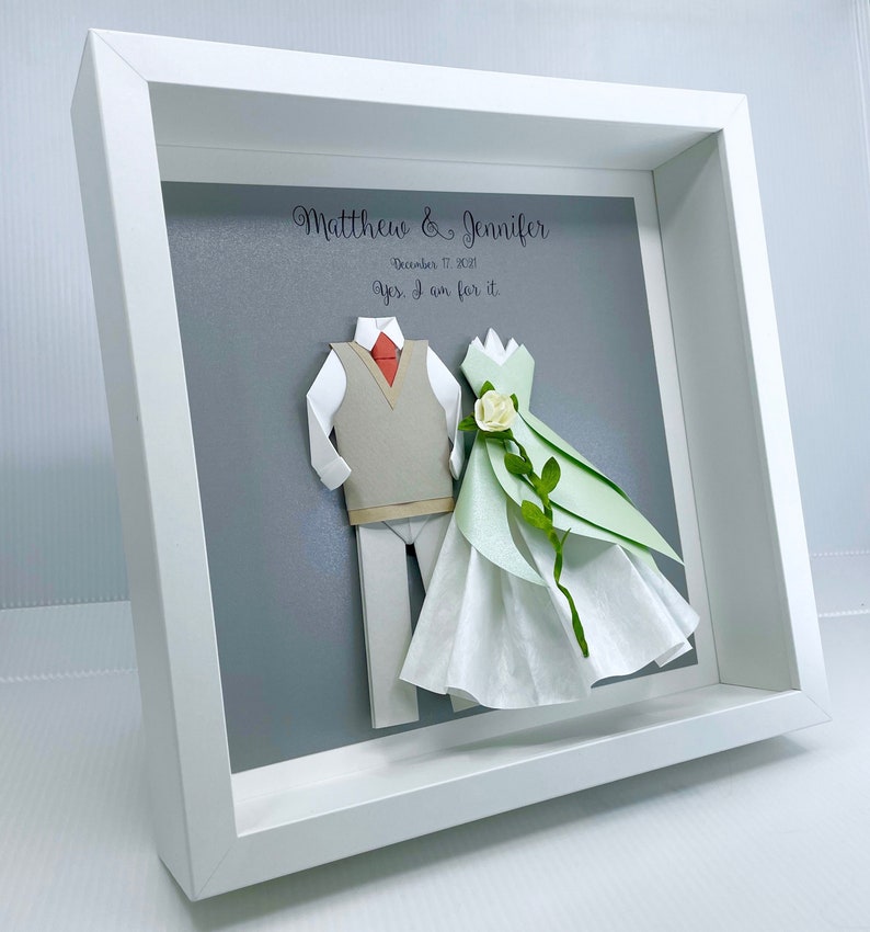 Peut inclure: Un cadre blanc avec un fond gris repr&eacute;sentant un couple en origami de papier. Le mari&eacute; porte un gilet gris et un pantalon blanc. La mari&eacute;e porte une robe verte et blanche avec une fleur blanche. Le texte "Matthew & Jennifer" et "December 17, 2021" est imprim&eacute; au-dessus du couple. Le texte "Yes, I am for it" est imprim&eacute; en dessous du couple.