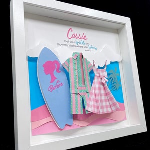 K&ouml;nnte beinhalten: Ein gerahmtes Papierbastelwerk mit einem blauen Surfbrett, einem pink- und gr&uuml;n gestreiften Hemd, einem pink- und wei&szlig; karierten Kleid und dem Text "Cassie Get your sparkle on. Show this world where you belong. -Anchia Barbie"