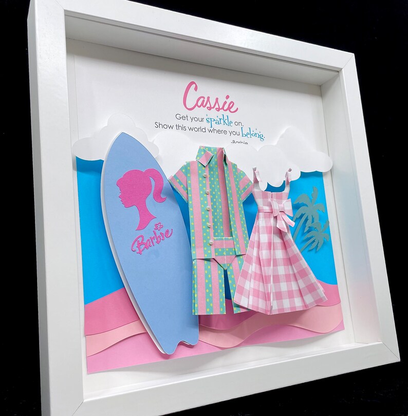 K&ouml;nnte beinhalten: Ein gerahmtes Papierbastelwerk mit einem blauen Surfbrett, einem pink- und gr&uuml;n gestreiften Hemd, einem pink- und wei&szlig; karierten Kleid und dem Text "Cassie Get your sparkle on. Show this world where you belong. -Anchia Barbie"