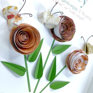 Pode incluir: Uma arte de papel com tr&ecirc;s flores de papel marrom e branco com folhas de papel verde e tr&ecirc;s borboletas com padr&otilde;es florais. As borboletas s&atilde;o feitas de papel e t&ecirc;m antenas de arame.