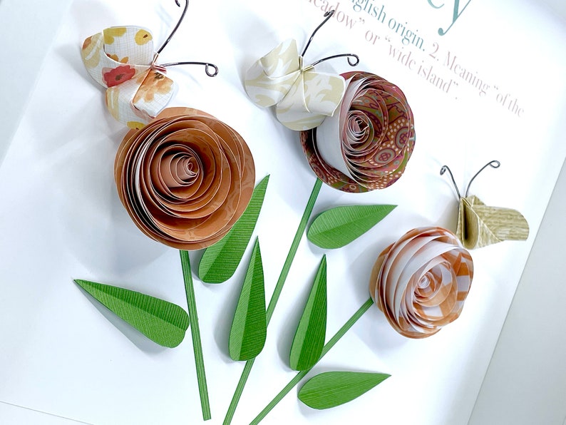 Pode incluir: Uma arte de papel com tr&ecirc;s flores de papel marrom e branco com folhas de papel verde e tr&ecirc;s borboletas com padr&otilde;es florais. As borboletas s&atilde;o feitas de papel e t&ecirc;m antenas de arame.