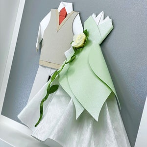 Peut inclure: Une cr&eacute;ation artisanale en papier encadr&eacute;e annon&ccedil;ant le mariage d'un couple. Le mari&eacute; porte un gilet gris et une cravate rouge. La mari&eacute;e porte une robe blanche avec un corsage vert et une fleur blanche. Le texte sur le cadre indique "December 17, 2021. Yes, I am for it."
