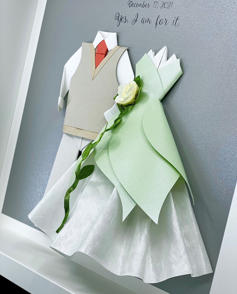 Peut inclure: Une cr&eacute;ation artisanale en papier encadr&eacute;e annon&ccedil;ant le mariage d'un couple. Le mari&eacute; porte un gilet gris et une cravate rouge. La mari&eacute;e porte une robe blanche avec un corsage vert et une fleur blanche. Le texte sur le cadre indique "December 17, 2021. Yes, I am for it."