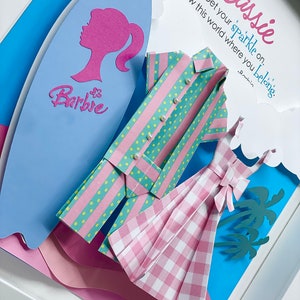 K&ouml;nnte beinhalten: Ein Papierbastelkunstwerk mit einem rosa-gr&uuml;nen gestreiften Hemd, einem rosa-wei&szlig;en karierten Kleid und einem blauen Surfbrett mit dem Wort "Barbie" darauf. Der Hintergrund ist blau mit rosa Wolken und Palmen. Der Text "Cassie Get your sparkle on. Show this world where you belong." ist in rosa und blau geschrieben.