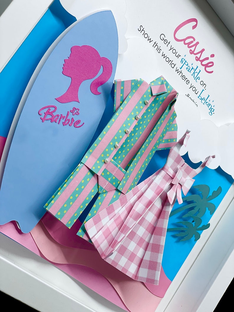 K&ouml;nnte beinhalten: Ein Papierbastelkunstwerk mit einem rosa-gr&uuml;nen gestreiften Hemd, einem rosa-wei&szlig;en karierten Kleid und einem blauen Surfbrett mit dem Wort "Barbie" darauf. Der Hintergrund ist blau mit rosa Wolken und Palmen. Der Text "Cassie Get your sparkle on. Show this world where you belong." ist in rosa und blau geschrieben.