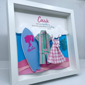 K&ouml;nnte beinhalten: Ein wei&szlig;er Rahmen mit einer blauen, rosa und gr&uuml;nen Papierbastel-Szene im Inneren. Die Szene enth&auml;lt ein Surfbrett mit einer rosa Silhouette eines Frauenkopfes und dem Wort "Barbie" darauf. Au&szlig;erdem gibt es ein rosa-gr&uuml;nes gestreiftes Hemd, ein rosa-wei&szlig;es kariertes Kleid und Palmen. Der Text "Cassie Get your sparkle on. Show this world where you belong. -Barbie" ist auf dem wei&szlig;en Hintergrund gedruckt.