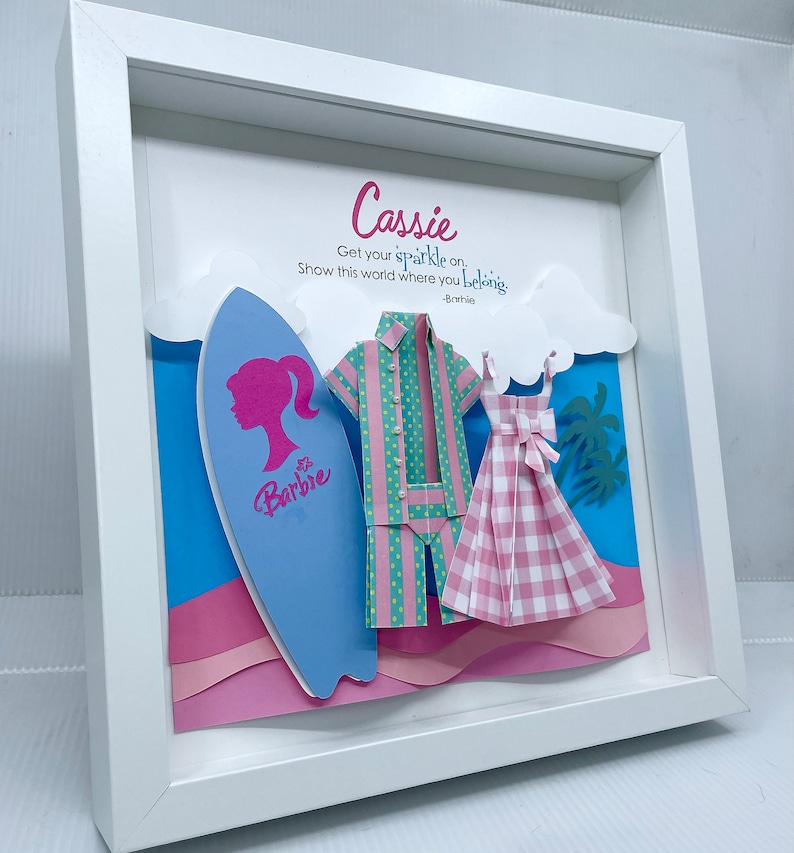 K&ouml;nnte beinhalten: Ein wei&szlig;er Rahmen mit einer blauen, rosa und gr&uuml;nen Papierbastel-Szene im Inneren. Die Szene enth&auml;lt ein Surfbrett mit einer rosa Silhouette eines Frauenkopfes und dem Wort "Barbie" darauf. Au&szlig;erdem gibt es ein rosa-gr&uuml;nes gestreiftes Hemd, ein rosa-wei&szlig;es kariertes Kleid und Palmen. Der Text "Cassie Get your sparkle on. Show this world where you belong. -Barbie" ist auf dem wei&szlig;en Hintergrund gedruckt.