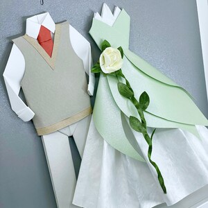 Peut inclure: Une &oelig;uvre d'art en papier encadr&eacute;e repr&eacute;sentant un couple habill&eacute; en origami. Le mari&eacute; porte une chemise blanche, un gilet gris et une cravate rouge. La mari&eacute;e porte une robe verte avec une rose blanche et des feuilles vertes. Le texte "December 17, 2021 Yes, I am for it." est &eacute;crit sur le fond gris.