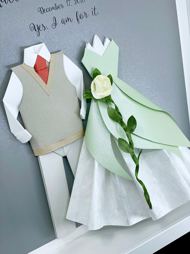 Peut inclure: Une &oelig;uvre d'art en papier encadr&eacute;e repr&eacute;sentant un couple habill&eacute; en origami. Le mari&eacute; porte une chemise blanche, un gilet gris et une cravate rouge. La mari&eacute;e porte une robe verte avec une rose blanche et des feuilles vertes. Le texte "December 17, 2021 Yes, I am for it." est &eacute;crit sur le fond gris.