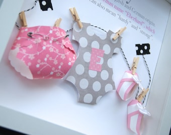 May include: A framed baby shower or nursery decor piece with a pink and white diaper, a gray and white polka dot onesie with the letter "E" on it, and two pink and white striped booties. The text in the frame reads "A man name "Eberhard", which is of English and German origin. It can also mean "hardy" and "strong."