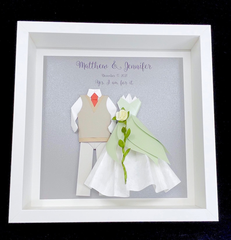 Peut inclure: Un cadre blanc avec un fond gris repr&eacute;sentant une d&eacute;coupe en papier d'un couple en tenue de mariage. Le texte "Matthew & Jennifer" et "December 17, 2021" est &eacute;crit en violet. Le texte "Yes, I am for it" est &eacute;crit en violet sous la date.
