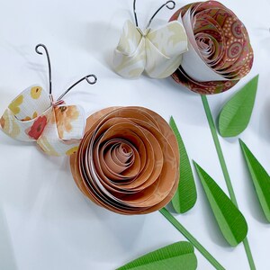 Pode incluir: Artesanato de papel com uma borboleta e flores. A borboleta &eacute; feita de papel branco e amarelo com um padr&atilde;o floral. As flores s&atilde;o feitas de papel marrom e laranja com um padr&atilde;o de espiral. As flores t&ecirc;m folhas de papel verde.