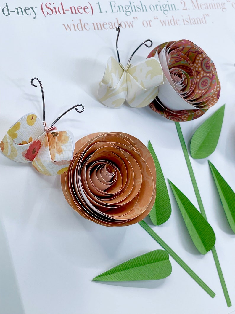 Pode incluir: Artesanato de papel com uma borboleta e flores. A borboleta &eacute; feita de papel branco e amarelo com um padr&atilde;o floral. As flores s&atilde;o feitas de papel marrom e laranja com um padr&atilde;o de espiral. As flores t&ecirc;m folhas de papel verde.