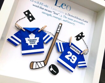 May include: A white shadow box frame containing two blue and white paper hockey jerseys, a miniature hockey stick, and a puck. One jersey features the Toronto Maple Leafs logo, while the other displays the name MARCHILDON and the number 29.