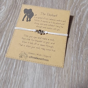 May include: A white string bracelet with a small silver elephant charm, displayed on a brown card. The card has an elephant silhouette and text about luck, patience, and wisdom. The text includes "Dream | Wish | Inspire" and a social media handle.