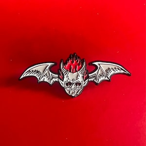 May include: A white enamel pin with a red and white devil head with wings. The devil has horns and a fiery red mohawk.