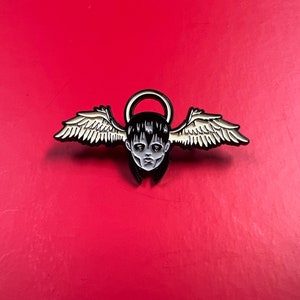 May include: Black and white enamel pin of a person with a halo and wings. The person has dark hair and a serious expression.