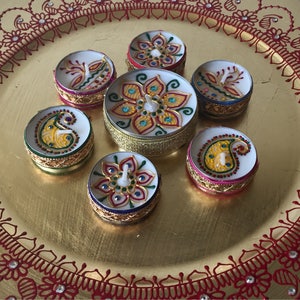 May include: A collection of seven decorative, round candles with intricate, colorful designs. The candles are arranged on a gold-colored plate with a red floral pattern around the edge. The candle designs include floral and paisley motifs.