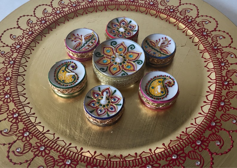 May include: A collection of seven decorative, round candles with intricate, colorful designs. The candles are arranged on a gold-colored plate with a red floral pattern around the edge. The candle designs include floral and paisley motifs.
