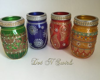 May include: Four glass jars decorated with intricate gold and silver henna-style designs. Each jar has a different colored base: green, blue, red, and yellow. The jars have a silver lid with a gold trim and a gold chain hanging from the lid.