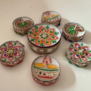 May include: A collection of seven decorative candles with intricate designs. The candles feature colorful floral and paisley patterns, with gold and silver metallic accents. The candles are round and have a white wax base.