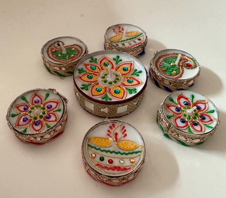 May include: A collection of seven decorative candles with intricate designs. The candles feature colorful floral and paisley patterns, with gold and silver metallic accents. The candles are round and have a white wax base.