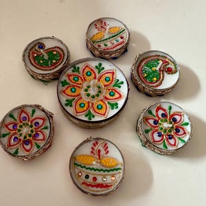May include: A collection of seven small, round decorative boxes with intricate designs. The boxes feature white backgrounds with colorful floral and paisley patterns, outlined in silver metal. The designs include red, green, yellow, and blue accents.