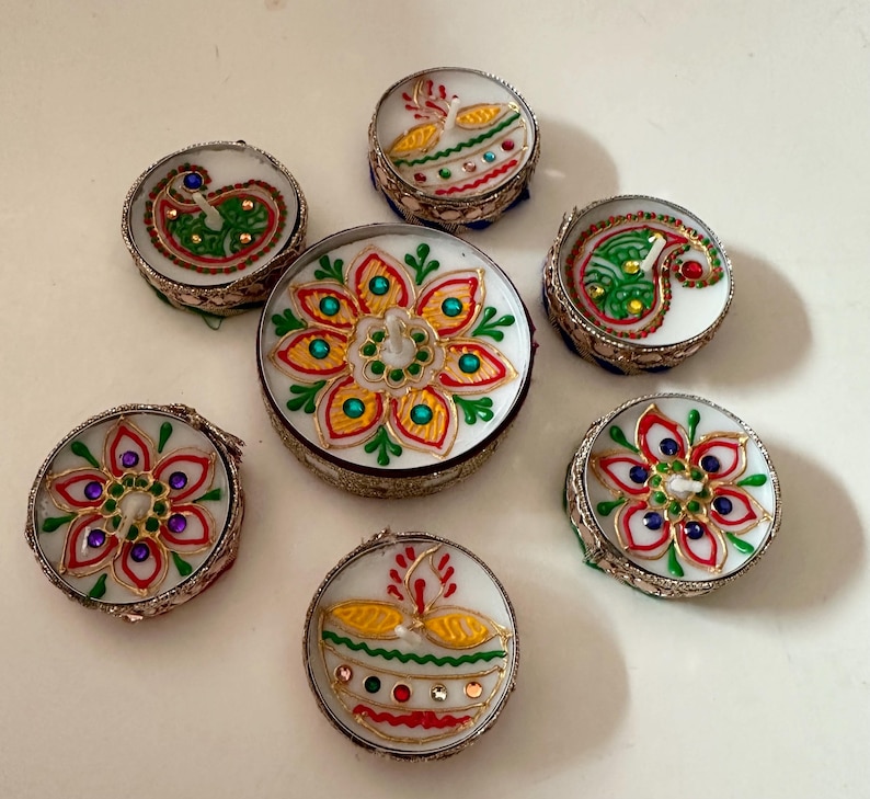 May include: A collection of seven small, round decorative boxes with intricate designs. The boxes feature white backgrounds with colorful floral and paisley patterns, outlined in silver metal. The designs include red, green, yellow, and blue accents.