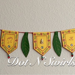 May include: A decorative banner with yellow pennants, each adorned with intricate designs and embellished with rhinestones. Green leaf cutouts and red string connect the pennants. The banner is suspended against a neutral wall.