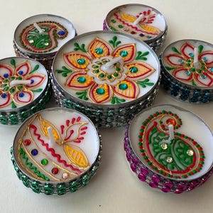 May include: Seven decorative tealight candles with intricate hand-painted designs. Each candle features a unique floral or paisley pattern in vibrant colors like red, green, and gold, accented with small gemstones. The candle holders are embellished with colorful jewels.