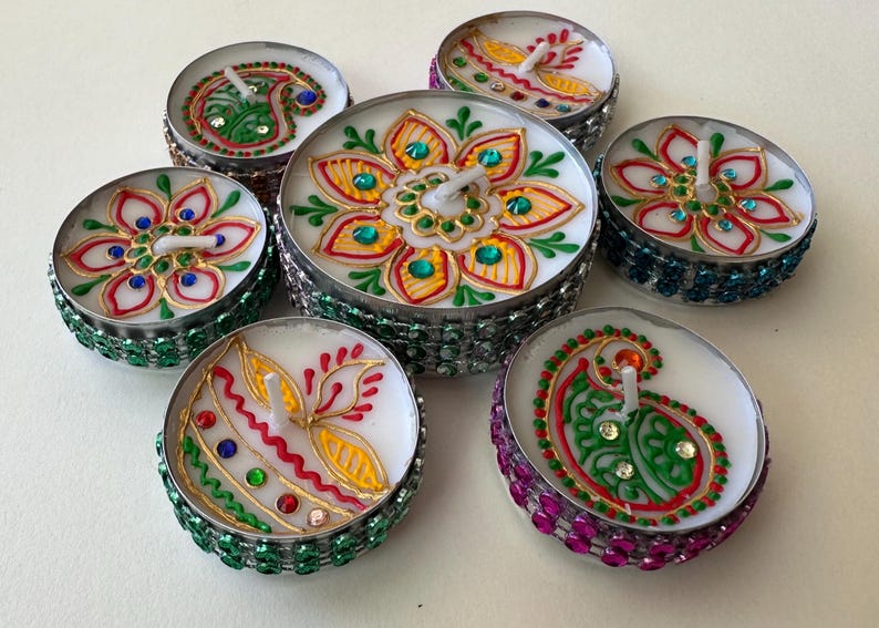 May include: Seven decorative tealight candles with intricate hand-painted designs. Each candle features a unique floral or paisley pattern in vibrant colors like red, green, and gold, accented with small gemstones. The candle holders are embellished with colorful jewels.