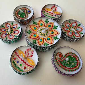 May include: A collection of seven decorative tea light candles. Each candle features a unique hand-painted design with vibrant colors, including red, green, yellow, and blue. The candles are set in silver-colored holders with decorative embellishments.