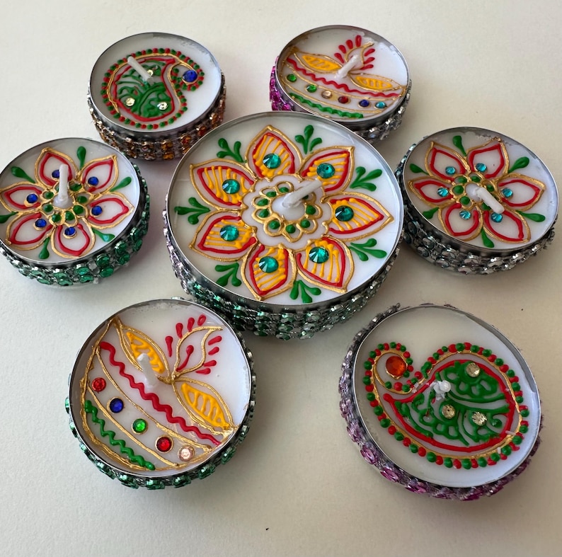 May include: A collection of seven decorative tea light candles. Each candle features a unique hand-painted design with vibrant colors, including red, green, yellow, and blue. The candles are set in silver-colored holders with decorative embellishments.