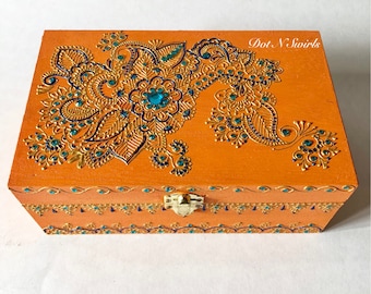 Puede incluir: Caja de madera naranja con un diseño paisley dorado y turquesa y diamantes de imitación. La caja tiene una tapa con bisagras y un cierre de metal. El texto "Dot N Swirls" está impreso en la parte superior de la caja.