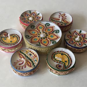 May include: A collection of seven decorative tea light candles. Each candle features a white wax base with a colorful, hand-painted design. The designs include floral and paisley patterns in gold, red, green, and blue, with decorative borders.