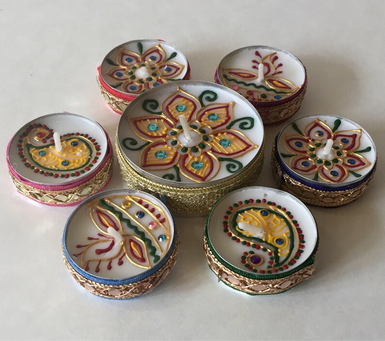 May include: A collection of seven decorative tea light candles. Each candle features a white wax base with a colorful, hand-painted design. The designs include floral and paisley patterns in gold, red, green, and blue, with decorative borders.
