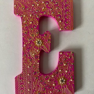 May include: A decorative wooden letter "E" in a vibrant pink hue, adorned with intricate gold filigree patterns. The design includes faux pearls and rhinestones, creating a luxurious and ornate appearance. The letter is a unique piece of home decor.