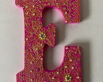 Puede incluir: Una letra "E" decorativa de madera en un vibrante color rosa, adornada con intrincados patrones de filigrana dorada. El diseño incluye perlas de imitación y pedrería, creando una apariencia lujosa y ornamentada. La letra es una pieza única de decoración del hogar.