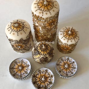 Puede incluir: Conjunto de seis velas blancas en forma de pilar con dise&ntilde;os de henna dorados y marrones y diamantes de imitaci&oacute;n. El conjunto incluye tres velas de pilar y tres velas de t&eacute;.