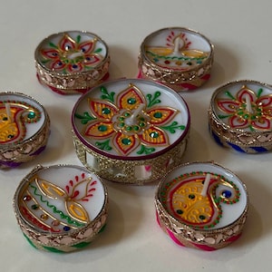 May include: A collection of decorative tealight candles with intricate designs. The candles feature colorful patterns, including floral and paisley motifs, with gold trim and jewel accents. The candles are arranged on a white surface.