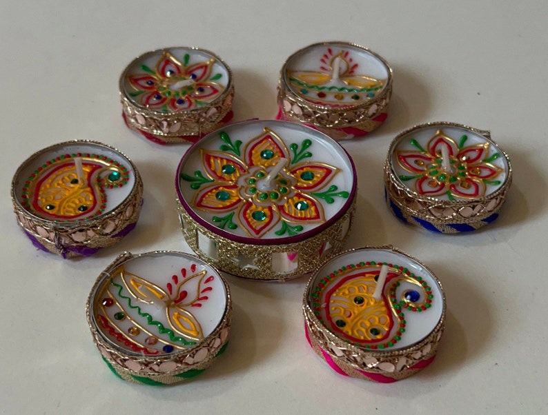 May include: A collection of decorative tealight candles with intricate designs. The candles feature colorful patterns, including floral and paisley motifs, with gold trim and jewel accents. The candles are arranged on a white surface.
