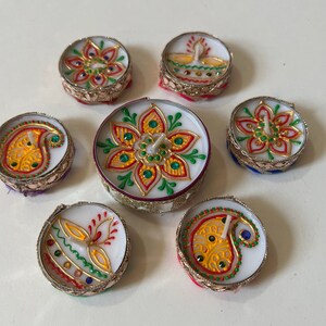 May include: A collection of seven decorative tea light candles. Each candle features a white wax base with colorful floral and paisley designs. The candles are adorned with gold trim and jewel accents, creating a festive and elegant appearance.