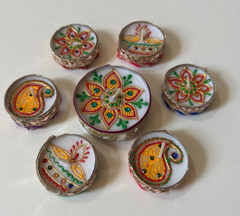 May include: A collection of seven decorative tea light candles. Each candle features a white wax base with colorful floral and paisley designs. The candles are adorned with gold trim and jewel accents, creating a festive and elegant appearance.